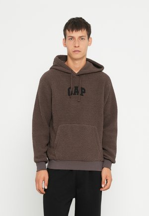 Hnedá flísová mikina s klokaním vreckom, ktorá obsahuje čierne logo "GAP". Mäkká textúra, nastaviteľná kapucňa so šnúrkami a ribbované manžety.