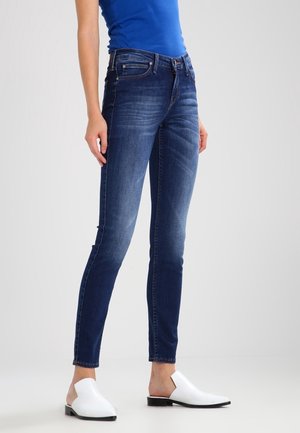 Vrouw draagt donkerblauwe skinny jeans, een blauw mouwloos topje en witte instappers, staand tegen een effen lichte achtergrond.