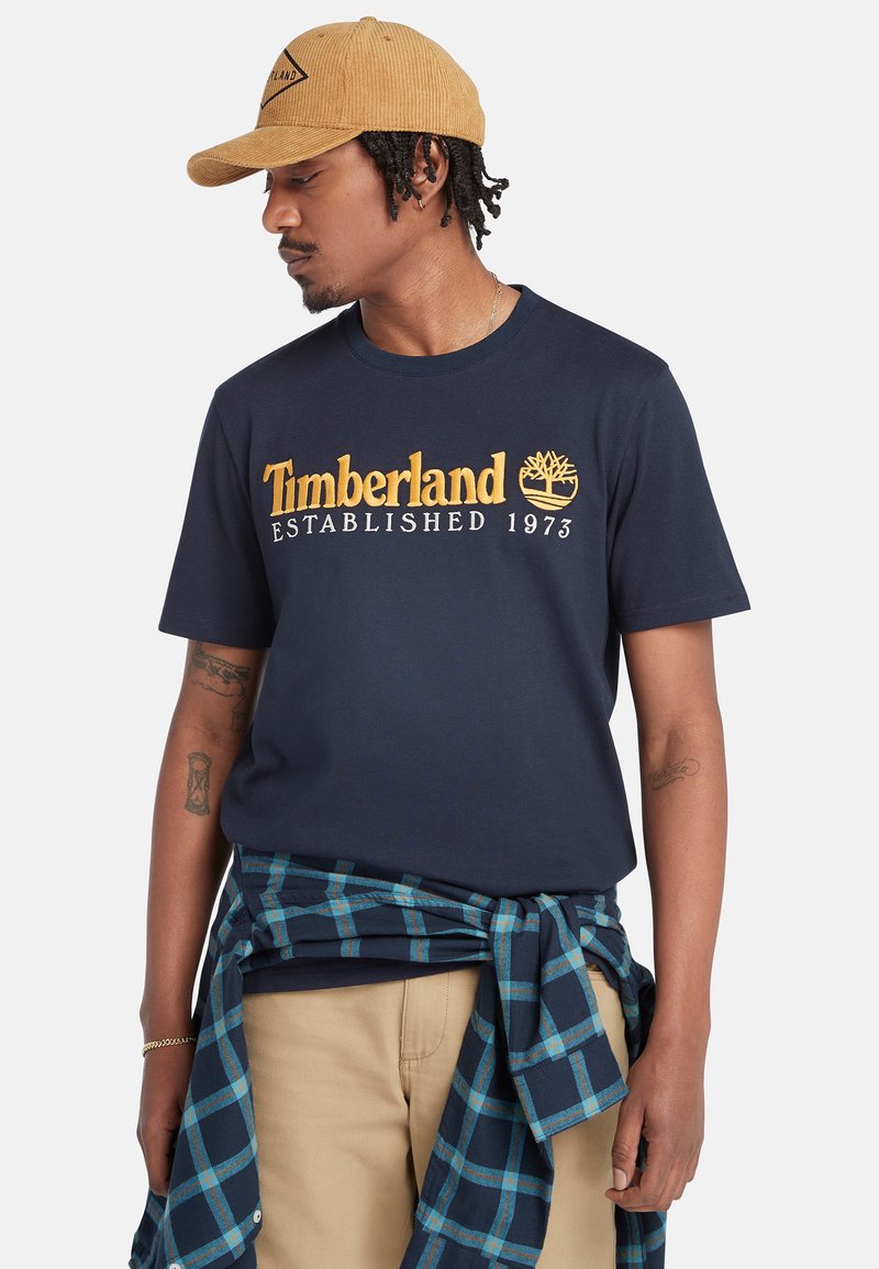 Námořnické tričko s žlutým textem a logem "Timberland Established 1973". Doplněno o modrou kostkovanou flanelovou košili uvázanou kolem pasu.