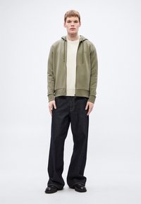 Felpa con zip color verde oliva sopra un maglione crema a coste, abbinata a jeans larghi in denim scuro e scarpe nere, in piedi davanti a uno sfondo neutro.