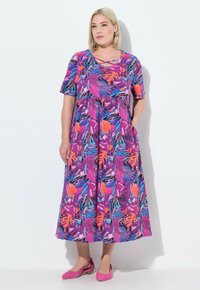 Lila maxi Kleid mit blumigem Muster, kurzen Ärmeln, gestuftem Design, Taschen und lebhaften tropischen Mustern, kombiniert mit rosa Ballerinas.
