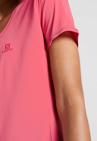 Short-sleeve, V-neck athletic shirt in bright pink, made of lightweight, textured fabric. Features a logo on the front.