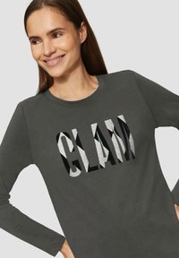 Donkergrijze shirt met lange mouwen, waarop het woord "GLAM" staat in een mix van glanzend zwart en glinsterend zilver lettertype. Zachte stof, ronde halslijn.