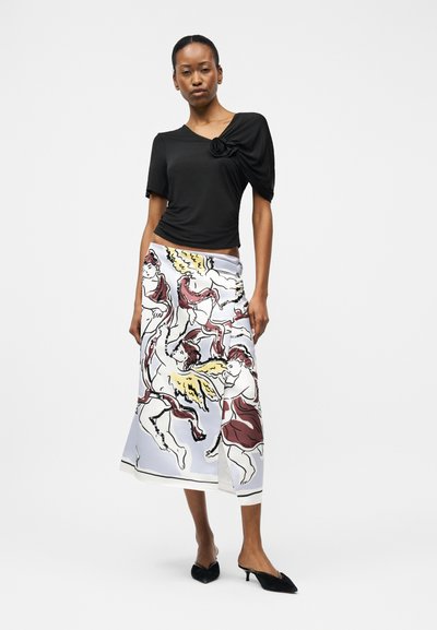 Γυναίκα στέκεται φορώντας μία μαύρη κοντομάνικη μπλούζα με λεπτομέρεια υφασμάτινου λουλουδιού και μία λευκή μίντι φούστα με σχέδιο αγγέλων και ερωτιδών.