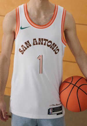 Personne tenant un ballon de basket portant un maillot blanc de San Antonio avec le numéro 1 et une bordure orange, debout à l'intérieur.