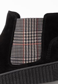 Bottines en suède noir avec un panneau latéral élastique à motif pied-de-poule et accents rouges. Semelle en caoutchouc noir texturé avec des stries.