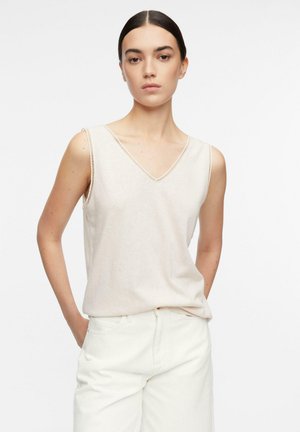 Kobieta o ciemnych włosach, ubrana w beżową bluzkę bez rękawów z dekoltem w kształcie litery V i białe spodnie, stoi ze złożonymi rękami w kieszeniach na jednolitym tle.