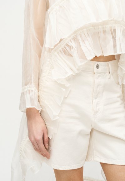 Κοντινό πλάνο ατόμου που φορά διάφανο λευκό μπλουζάκι με βολάν και μακριά μανίκια, σε συνδυασμό με λευκά ψηλόμεσα σορτς από τζιν.