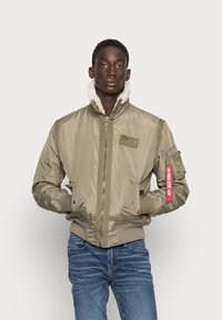 Olivgrön bomberjacka med en faux shearling-krage, dragkedjestängning och en röd taggdetalj. Har ribbade muddarna och sidofickor.