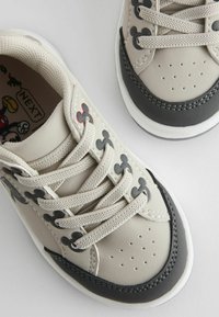 Beige and black children's sneakers with textured rubber, perforated toe, and gray laces featuring small Mickey Mouse accents.