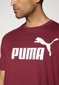 Bordeaux kortærmet T-shirt lavet af bomuld, med et fremtrædende hvidt "PUMA" logo og en springende puma grafik. Glat tekstur.