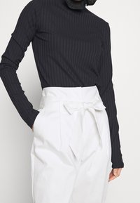 Zwarte geribbelde langemouwen top met hoge hals, gecombineerd met hoge witte broek met een strikceintuur en zijzakken.