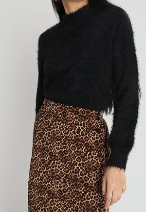 Kvinde iført en fluffy sort langærmet crop sweater og et højtaljet nederdel med leopardprint og sorte knapper i siden.