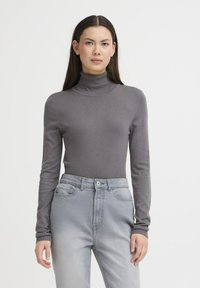 Pelēks garo piedurkņu augsto kaklu topiņš no gludas adīta materiāla, apvienots ar gaiši pelēkiem augstajā jostasvietā džinsiem, kuriem ir priekšējās kabatas un pogu aizdare.