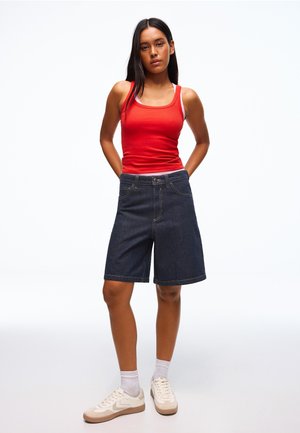 Jonge vrouw staat met handen achter haar rug, draagt een rood tanktopje, donkere denim korte broek tot aan de knieën, witte sokken en beige sneakers.