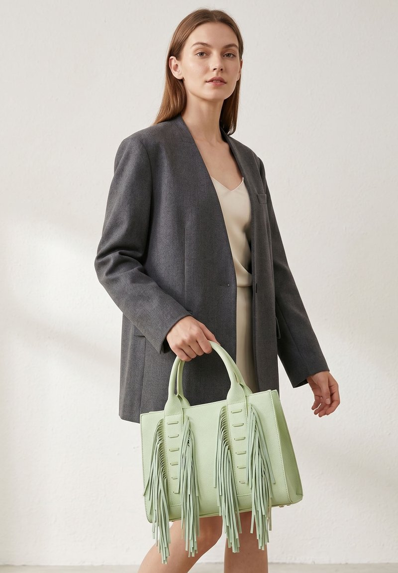 Femme portant un blazer gris sur une robe crème tient un sac à main vert clair avec de longs pompons à franges dans un espace neutre.