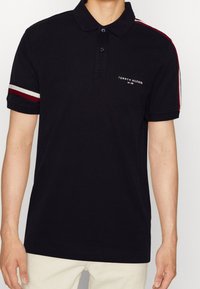 Černé polo tričko s límečkem, které má modré knoflíky, bílé a červené pruhované detaily na rukávech a malou nášivku na hrudi.