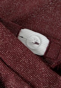 Burgunderfarget stoff med metalltråder brettet over et hvitt elastisk bånd festet med en klar, firhulls knapp.