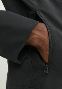 Zwarte jas met een ritszak op de mouw. De stof heeft een gladde textuur met een matte afwerking.
