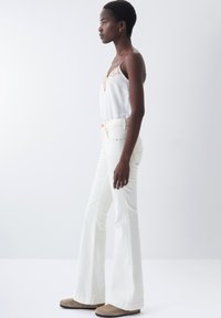 Jeans blancos acampanados combinados con una blusa de seda ligera con acentos de encaje, mostrados en un modelo de perfil contra un fondo liso.