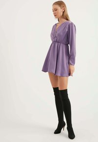 Lila Minikleid mit langen Ärmeln, V-Ausschnitt, gerafftem Bund und glattem Stoff. Kombiniert mit schwarzen Overknee-Stiefeln mit spitzen Zehen.