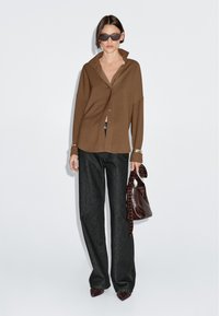 Brown oversized button-up shirt paired with dark wide-leg jeans. Accessorized with a brown handbag and sunglasses. Polished, modern design.