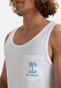 Camiseta sin mangas de algodón blanca con un bolsillo en el pecho y un gráfico azul de una palmera con un ojo, con la marca "BILLA BONG" debajo.