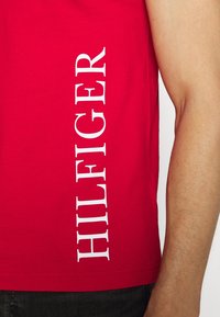Červený bavlněný top bez rukávů s vertikálním bílým textem, který říká "HILFIGER" na levé straně. látka vypadá hladce a lehce.