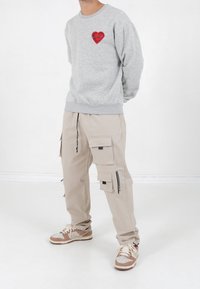 Grijze sweatshirt met rood hartembleem, gecombineerd met beige cargobroek met meerdere zakken en een zwarte ritsdetail, gedragen met bruin sneakers.