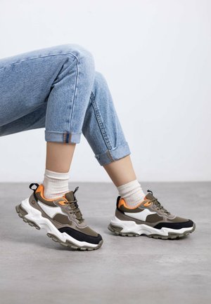 Zapatillas deportivas en verde oliva, blanco y negro con detalles en naranja. Suela gruesa, materiales de malla y sintéticos, y suela de goma para tracción.