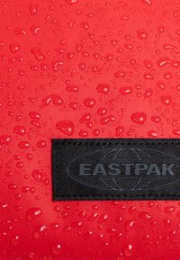 Tessuto rosso testurizzato con gocce d'acqua visibili, dotato di una patch nera del logo Eastpak in basso. Materiale resistente e impermeabile.