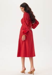 Abito rosso a maniche lunghe con vita arricciata e maniche a sbuffo. Tessuto liscio e leggero; lunghezza al ginocchio. Indossato con sandali beige con tacco.
