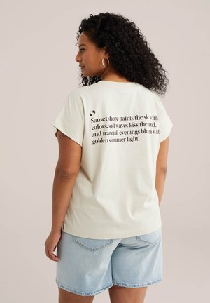 Femme aux cheveux bouclés portant un t-shirt crème avec un texte sur le thème du coucher de soleil et un short en jean bleu clair, debout devant un fond uni.