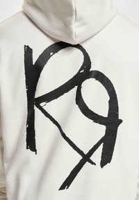 Hvid hættetrøje med et stort sort grafikdesign på ryggen. Materialet ser blødt ud, og pasformen inkluderer en hætte.