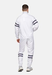 Witte trainingsoutfit met marineblauwe strepen bij de mouwen en onderkant, voorzien van een hoge kraag en elastische tailleband op de broek. Gemaakt van een gladde stof.