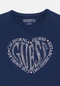 Mørkeblå T-skjorte i økologisk bomull med hjertedesign laget av rhinestones; teksten inkluderer "GUESS", "California" og "Los Angeles 1981".