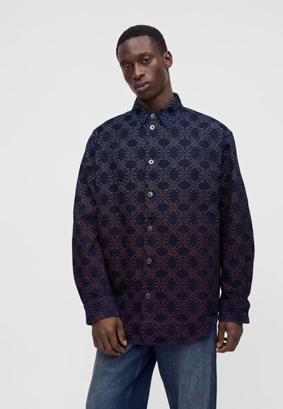 Jeune homme portant une chemise boutonnée bleu foncé à motifs et un jean bleu, debout devant un fond clair uni.