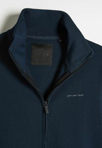 Granatowa kurtka zapinana na zamek z wysokim kołnierzem, miękką strukturą i czarną metką z białym nadrukiem. Zamek błyskawiczny biegnie wzdłuż centrum.