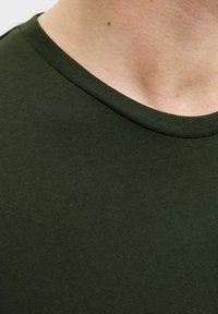 Detailní záběr na krk a horní část hrudi osoby oblečené v tmavě zeleném tričku s kulatým výstřihem.