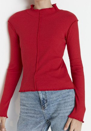 Personne portant un haut rouge côtelé à manches longues avec un bord volanté et un jean taille haute bleu clair sur un fond uni.