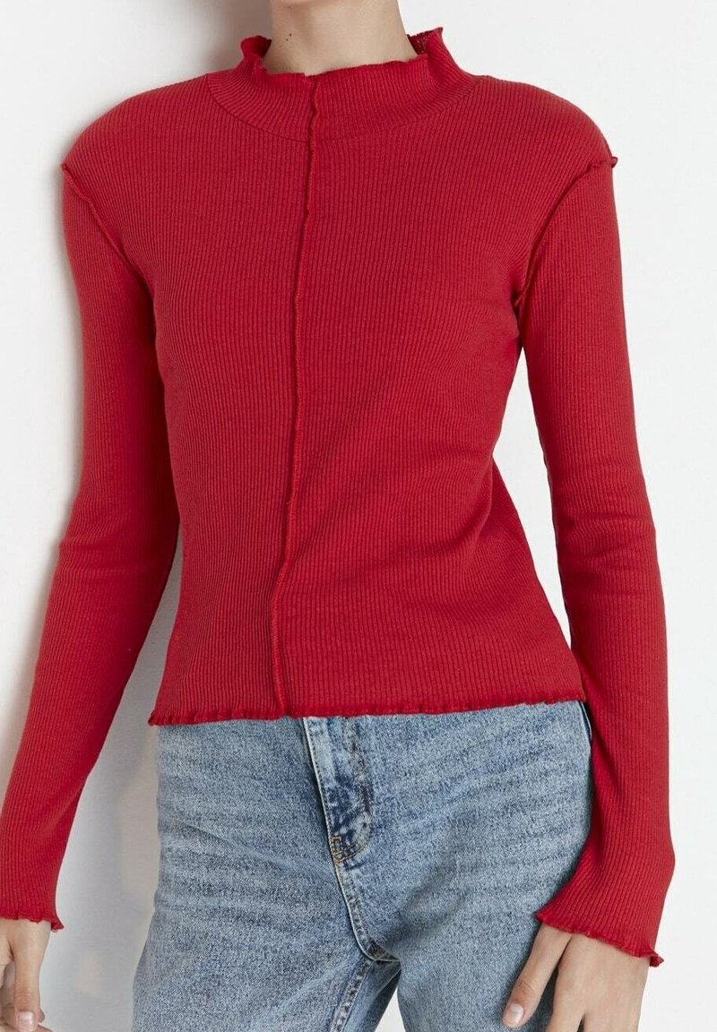 Personne portant un haut rouge côtelé à manches longues avec un bord volanté et un jean taille haute bleu clair sur un fond uni.