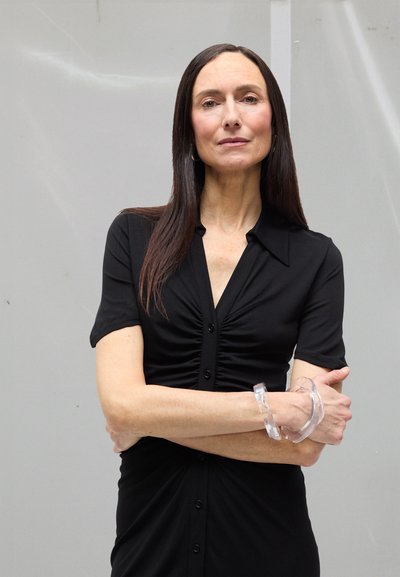 Vrouw met lang donker haar, gekleed in een zwarte jurk met knopen en een doorzichtige, dikke armband, staand met gekruiste armen voor een effen grijze achtergrond.