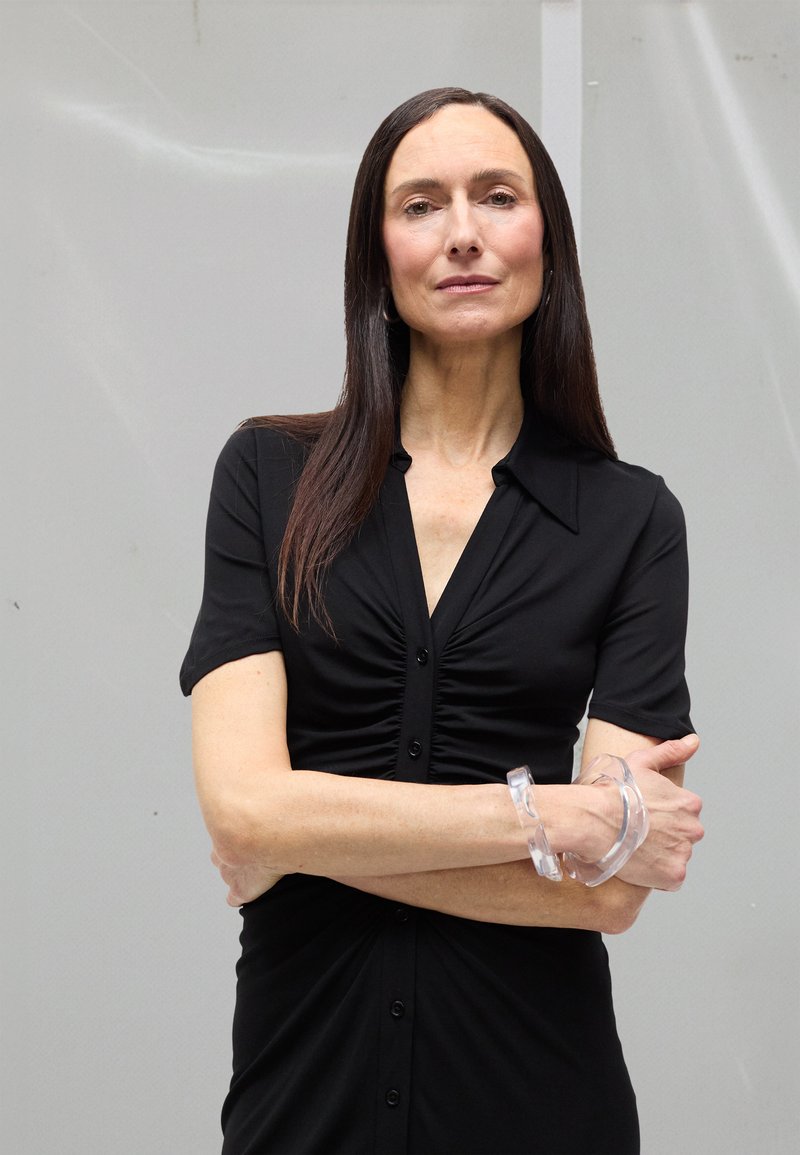 Woman with long dark hair wearing black buttoned dress, transparent chunky bracelet, standing with arms crossed against plain gray background.