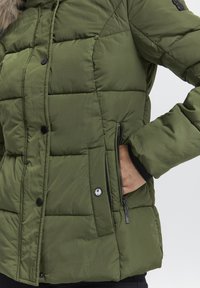 Olivengrøn pufferjakke med quiltet mønster, frontknapper med trykknap og en lynlåslomme i siden. Har en blød tekstureret overflade.