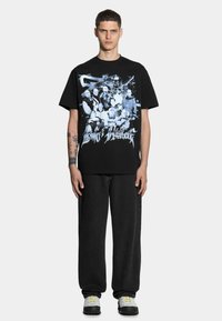 Czarna bawełniana koszulka z graficznym nadrukiem w odcieniu niebieskim, przedstawiającym wiele postaci. Połączona ze luźnymi czarnymi dżinsami i szarymi trampkami.