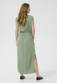Maxi dress in light green fabric, featuring short sleeves, an elastic waist, and side slit details. Worn with black flats.