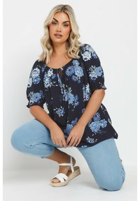 Blusa floral azul con escote redondeado y detalle de lazo; mangas cortas; combinada con jeans azul claro y sandalias de esparto blancas.