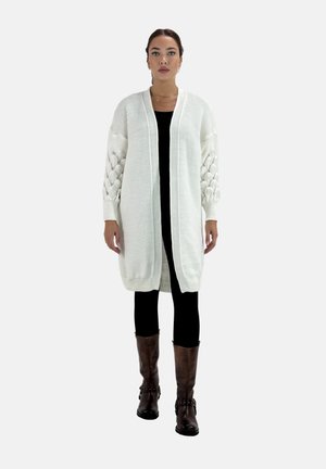 Cárdigan blanco de punto con frente abierto, mangas globo texturizadas y diseño acanalado. Combinado con leggings negros y botas marrones hasta la rodilla.