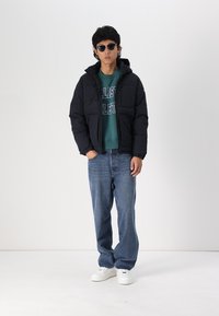 Sort dunjakke med hætte, teal sweatshirt med tekst, blå jeans og hvide sneakers. Tilbehør inkluderer sorte solbriller.