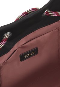 Interior de satén rosa de un bolso con un exterior negro, que cuenta con un asa trenzada con acentos en rosa, negro y crema, y una etiqueta negra con "TOUS."